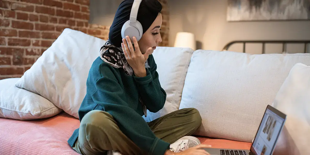 A female adult student, wearing a head scarf and a green loose jumper and trousers, sat cross legged on a sofa, with headphones on and working on a laptop.