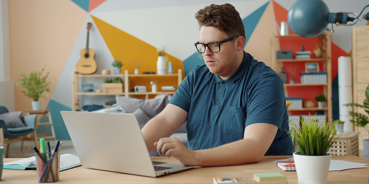 A male adult remote student, wearing glasses and a blue t-shirt, sat at a desk working on a laptop